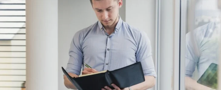 Man writing in a notebook while standing by a window.